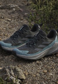 Schwarze und türkisfarbene Sportschuhe mit einer strukturierten Oberseite, Gore-Tex-Branding und grauen Akzenten, auf einer Kiesoberfläche platziert.