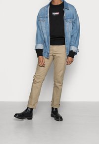 Giacca di denim, felpa nera con logo, pantaloni kaki e stivaletti neri alla caviglia. Caratteristiche includono maniche arrotolate e vestibilità casual.