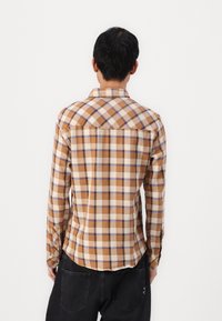 Man in een bruin, beige en blauwe geruite overhemd met lange mouwen en zwarte broeken, staand met zijn rug naar de camera tegen een witte achtergrond.