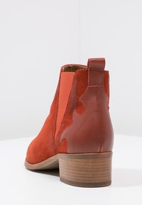 Botín de ante rojo con detalles en cuero contrastante, puntera redonda, tacón trasero corto y paneles elásticos laterales. Textura suave, diseño versátil.