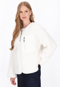 Weiße Fleecejacke mit weicher Textur, ausgestattet mit einer vorderen Reißverschlusstasche, langen Ärmeln und lockerer Passform. Trägt man über einem weißen Oberteil.