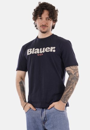 Uomo con capelli ricci e orecchini che indossa una maglietta blu navy "Blauer USA" e jeans azzurri, con le mani nelle tasche, mostrando tatuaggi su entrambe le braccia.