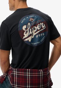 Camiseta negra con un gran gráfico circular envejecido en azul y burdeos, con texto y acentos en varios idiomas. Camisa de cuadros atada a la cintura.