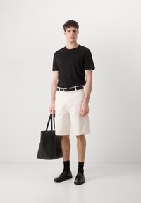 Schwarzes T-Shirt mit kurzen Ärmeln, cremefarbene Shorts, schwarzer Gürtel mit weißen Akzenten, schwarze Lederschuhe, schwarze Tragetasche mit glatter Textur.