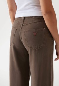 Pantalones de mezclilla marrones con un ajuste relajado, que cuentan con bolsillos traseros y una etiqueta triangular roja en el bolsillo derecho.