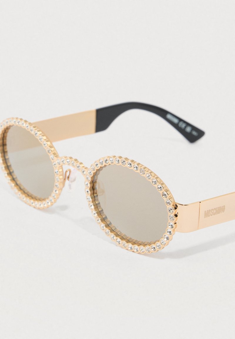 Goudkleurige zonnebril met ronde lenzen, omlijst met heldere strasstenen, voorzien van een transparant montuur en zwarte pootjes.