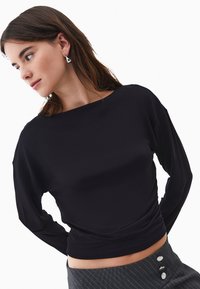 Ung kvinna med brunt hår bär silverfärgade triangelformade örhängen, en figursydd svart långärmad topp och grå rutiga byxor med hög midja och knappar.