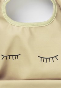 Bavaglino in silicone di colore beige pallido con scollo rotondo, caratterizzato da un design con occhi chiusi neri. Texture liscia con bordi cuciti.