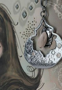 Srebrny metalowy wisiorek o teksturowej powierzchni, z wycięciami i jasnoróżowym koralikem. Przymocowany do łańcuszka na wzorzystym tle.