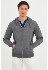 Sudadera gris con cremallera y acabado texturizado, dos bolsillos frontales y capucha con cordones. Lleva encima una camiseta de cuello en V de color azul marino.