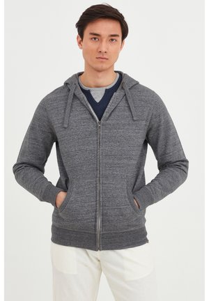Sudadera gris con cremallera y acabado texturizado, dos bolsillos frontales y capucha con cordones. Lleva encima una camiseta de cuello en V de color azul marino.