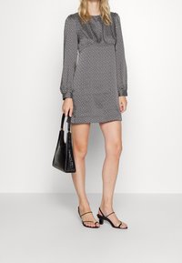 Vestido de manga larga con un patrón en blanco y negro, de corte holgado y cuello fruncido, combinado con un bolso negro brillante y tacones negros con tiras.