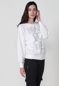 Felpa bianca con "BRAVE ♥ FREE" in strass, colletto a giro, maniche lunghe e vita elasticizzata. Abbinata a pantaloni cargo neri.