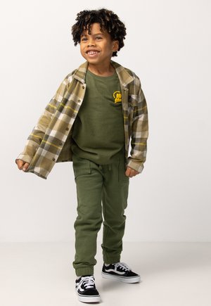 Lachend kind met krullend haar in een groene outfit en een open beige geruite shirt, staand met één arm iets omhoog.