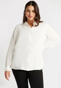 Biała koszula zapinana na guziki z długimi rękawami, zebranymi mankietami oraz luźnym krojem. Gładki materiał z lekkim połyskiem i zaokrąglonym dołem.