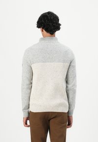Man met krullend, donker haar, gekleed in een tweekleurige gebreide trui in lichtgrijs en crème, gecombineerd met een paarse broek, die van zich af kijkt tegen een witte achtergrond.