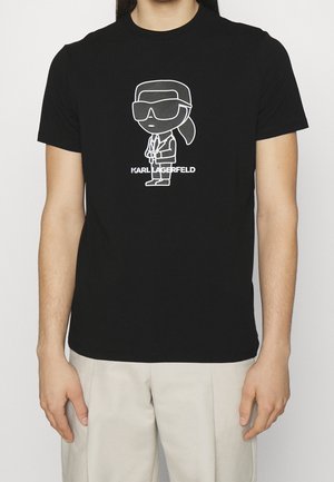 Osoba nosząca czarny t-shirt z białą kreskówkową postacią Karla Lagerfelda i tekstem "KARL LAGERFELD" z przodu.
