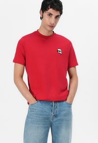 Raudona medvilninė marškinė su trumpomis rankovėmis ir apvaliu iškirpimu, su mažu juodai baltu grafiniu užkliju ant krūtinės.