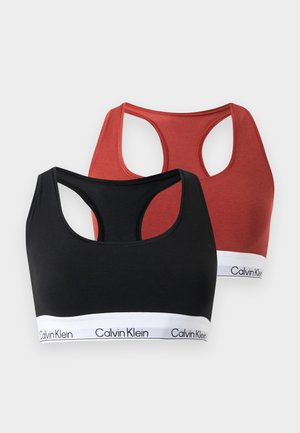 Femme debout pieds nus portant un soutien-gorge de sport Calvin Klein rouge et un slip assorti avec des bandes élastiques blanches à logo sur fond uni.