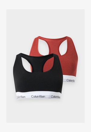 Femme debout pieds nus portant un soutien-gorge de sport Calvin Klein rouge et un slip assorti avec des bandes élastiques blanches à logo sur fond uni.