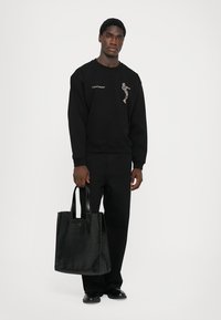 Felpa nera con dettagli ricamati, abbinata a pantaloni neri e una borsa tote nera lucida, con manici rettangolari.