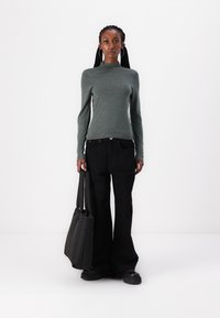 Grønn langermet topp med høy hals, svarte vide bukser og en svart totebag. Modellen står mot en hvit bakgrunn.