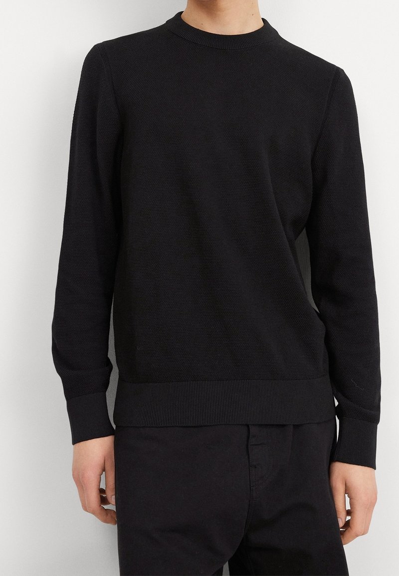 Černý pletený svetr s kulatým výstřihem z texturované látky, s žebrovanými manžetami a lemem. Přiléhavý střih, nošený s odpovídajícími černými kalhotami.