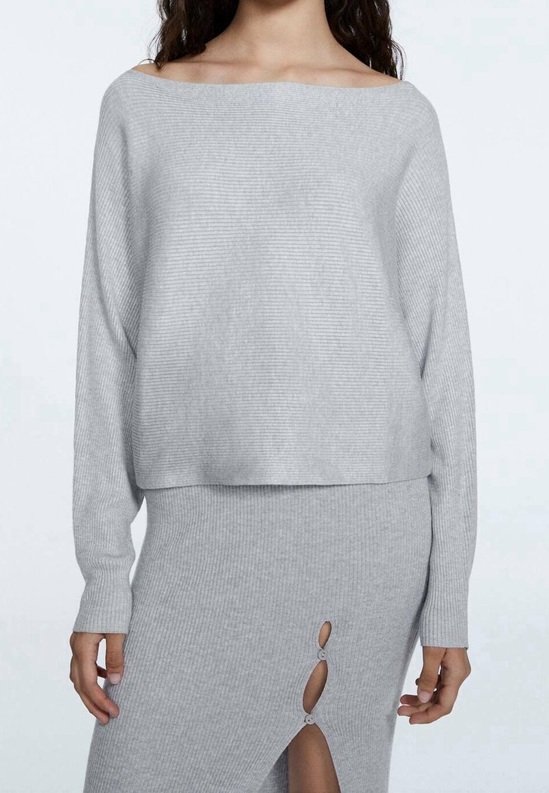 Femme portant un pull en maille côtelée gris clair et une jupe assortie avec deux découpes à boutons sur le devant et une fente.