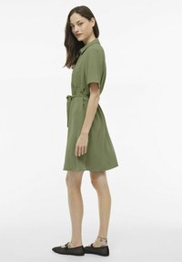 Alyvos žalios spalvos suknelė su trumpomis rankovėmis, apykakle ir diržo raiščiu. Pagaminta iš tekstūruoto audinio, turi priekinį kišenę. Dėvima su juodais butais.