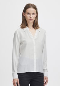Blusa bianca a maniche lunghe con colletto. Tessuto semi-trasparente, chiusura con bottoni e taglio dritto. Indossata con pantaloni neri.