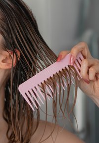 Różowy plastikowy grzebień o szerokich zębach, trzymany w dłoni, rozczesujący mokre brązowe włosy, z widocznymi kroplami wody i błyszczącą teksturą.