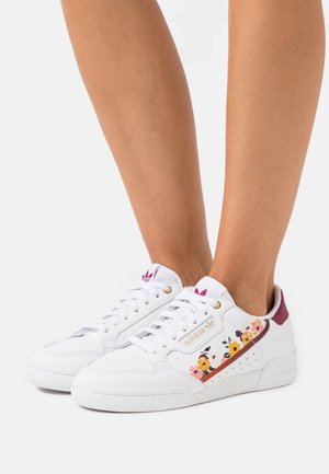 Baskets blanches Adidas avec motif floral sur le côté et accents bordeaux portées sur jambes nues devant un fond uni.
