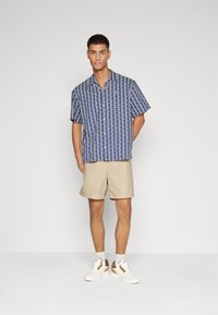 Blå kortärmad skjorta med geometriskt mönster, beiga shorts och vita sneakers med bruna och gröna accenter.