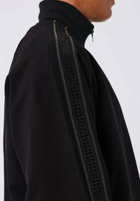 Giacca nera con zip, dotata di un colletto alto, pannello testurizzato nero lungo la spalla con accenti verticali e un design a vestibilità rilassata.