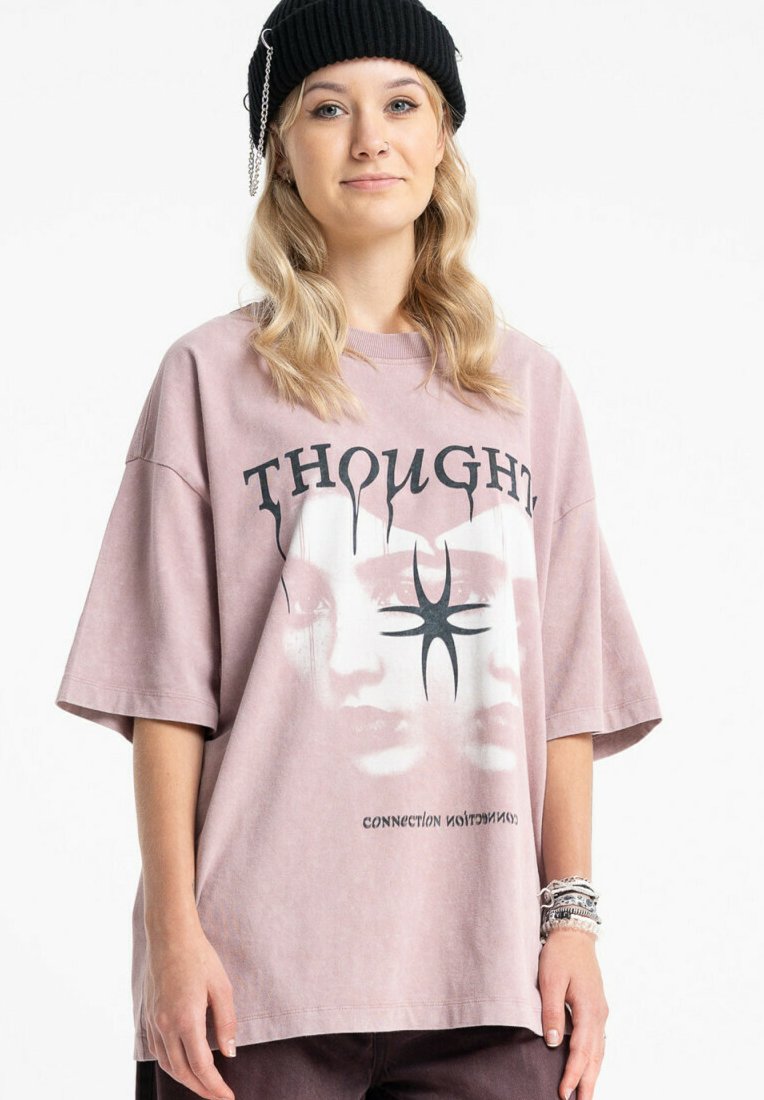 Luźna, krótkorękawowa koszulka w kolorze kurzowego różu, wykonana z bawełny, z graficznym wzorem przedstawiającym twarze i napisem "THOUGHT". Napis jest czarny, krój swobodny.
