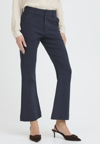 Pantalones de campana en tejido ligero, en color azul marino, con pliegues frontales, una cintura de altura media y un dobladillo limpio. Combinados con tacones de cuero puntiagudos.