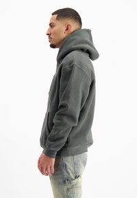 Donkergroene hoodie van zachte stof, met een kangoeroezak, ribbelboorden en een opvallende capuchon. Draag het met lichte denim broek.