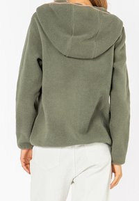 Persona vestida con un hoodie de forro polar verde oliva y pantalones blancos, vista desde atrás sobre un fondo blanco.