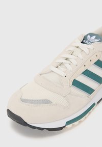 Beige sneaker met teal accenten, bovenwerk van suede en mesh, witte veters en een rubberen zool. Voorzien van een drie-strepen logo en een gevoerde kraag.