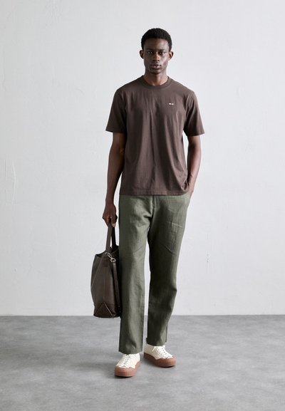 T-shirt marron foncé, pantalon en lin vert, baskets blanches avec des rayures brunes, portant un sac à épaule gris. Design simple, coupe décontractée.