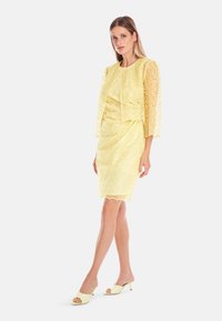 Gelbes Spitzenkleid mit Dreiviertelärmel, taillierter Silhouette und gewelltem Saum; getragen mit hellgelben offenen High Heels.