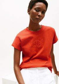 Czerwona bawełniana koszulka z podwiniętymi rękawami, z haftowanym logo na środku. Gładka faktura, luźny krój. W zestawieniu z białymi spodniami.