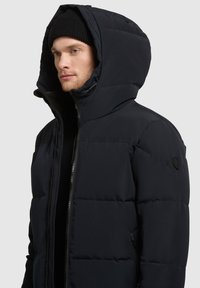 Chaqueta acolchada de color azul oscuro con un alto cuello, gran capucha, cierre de cremallera y patrón de costura horizontal. Presenta un logo en la manga.