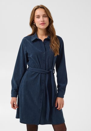 Femme aux longs cheveux bruns portant une robe bleu marine boutonnée jusqu'aux genoux, à manches longues et une ceinture à nouer à la taille, debout devant un fond uni.