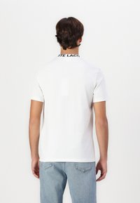 T-shirt blanc à manches courtes avec un col ras du cou, présentant un logo noir sur le col. Tissu lisse, coupe décontractée et dos uni.