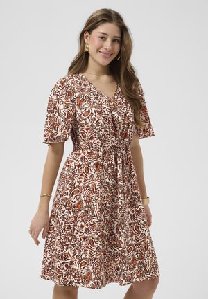 Kvinde med langt brunt hår iført guldsmykker i ørerne og armbånd, smilende i en knælængde kjole med brune og orange blomsterprint.