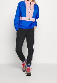 Blå og pink vindjakke med frontlynlås, elastiske manchetter og et adidas-logo. Sorte joggers med hvide striber på siderne og klassiske sneakers.