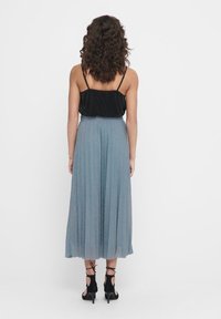 Top nero senza maniche abbinato a una lunga gonna plissettata in blu metallico e sandali con tacco neri, visti di spalle.