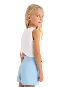 Top bianco senza maniche con uno spacco posteriore, abbinato a pantaloni corti azzurri chiari con una texture liscia e una cintura. Ragazza con lunghi capelli biondi.