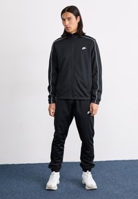 Une personne avec de longs cheveux porte un survêtement noir Nike et des baskets blanches, se tenant sur un tapis bleu avec un mur blanc uni en arrière-plan.